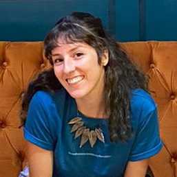 Foto de uma jovem mulher branca de cabelos castanhos cacheados e franja. Sentada em um sofá, ela sorri e faz contato visual. Ela veste uma blusa azul e o sofá laranja contrasta com uma parede também azul.